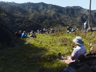 Ramble Worldwide walking group sat resting on grass enjoying lunch and conversation with mountain view in distance, La Gomera, Canary Islands, Spain