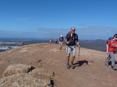 Man and woman walking atop peak wearing hiking boots and using trekking poles on sunny day in Lanzarote with rest of Ramble Worldwide walking group behind them and vast mountainous landscape, Canary Islands, Spain