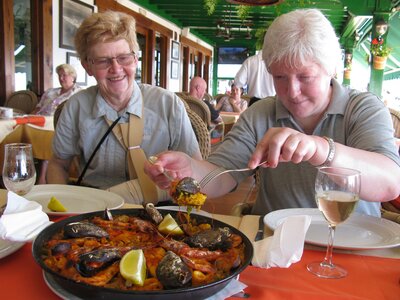 Close-up of two women at restaurant dining table excited by flavourful Spanish paella dish with seafood and lemon wedges on top, one moving food to their plate with a smile on their face, Lanzarote, Spain