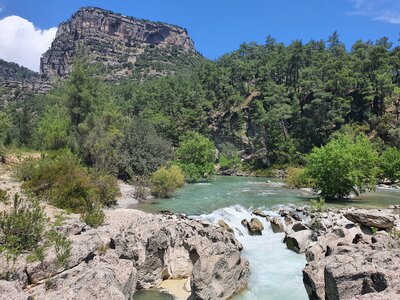 Tall rocky peak and hills covered in green shurbs and pine trees in distance with body of water flowing towards foreground at Koprulu Canyon National Park on sunny day, Turkey