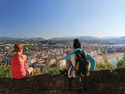 Two women sat on wall taking in view of city, beach, and mountains in distance whilst on the walking trail of Way of St James, Camino de Santiago