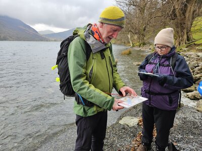 Man and woman on Ramble Worldwide Navigation & Hill Skills course reading maps with compasses in Lake District on shore of Buttermere Lake