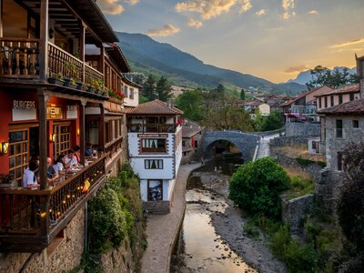 People sat on balcony next to rio quiviesa river in town of Potes with sunset in distance amidst mountains, Potes town, Cantabria, Spain