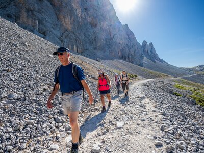 Ramble Worldwide walking holiday group descending long gradual rocky path in Picos De Europa mountain range with sun shining above mountain in background, Spain