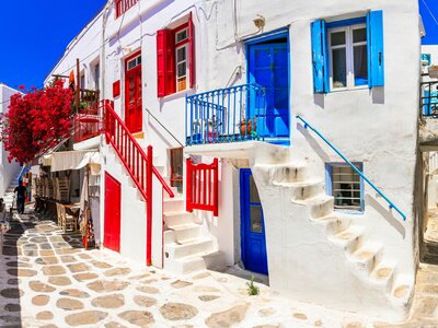 Colourful blue and red painted doors and windows in street of old Chora village with red flowering plant growing in distance arching over stone pattern pathway, Ios, Cyclades, Greece