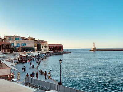 Spaced-out groups of people walking along a pier next to the ocean with lighthouse in distance, Chania, Crete, Greece