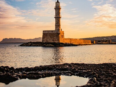 Lighthouse near body of water during sunset with reflection op top of lighthouse in small puddle on pebble shore, Chania, Greece