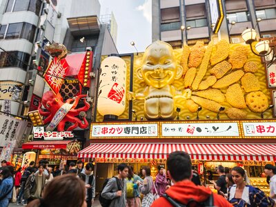 Crowds of people walking around outside front of Japanese street vendor shops and restaurants with colourful display of large octopus and golden baby holding food, Dotonbori street in Osaka, Japan