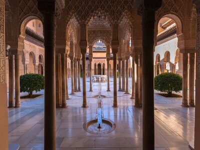 Decorated beige pillars with marble flooring reflecting sunlight from outdoor garden space in Alhambra palace, Granada, Spain