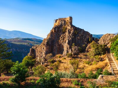 Lanjaron Castle atop tall rocky peak with mountains in far distance on sunny day, Granada, Spain