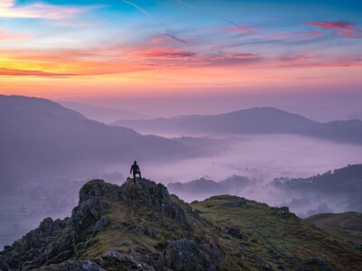 Hiker in distance standing on rocky peak admiring beautiful Lake District orange sunrise from a summit over the misty valley of Grasmere