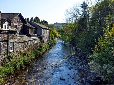 Stream running through Grasmere  next to traditional stone built houses and tall green trees with sun shining through, Lake District in Cumbria, UK