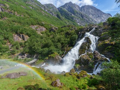 Beautiful large waterfall with rainbow created from mist on sunny day in mountainous green landscape, Briksdalsbreen waterfall in Norway