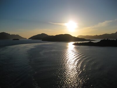 Cruise trail and low-hanging sun beaming golden ray amongst Norwegian water with small island silhouettes in far distance