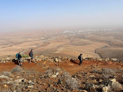 Side angle view of trio of walkers descending orange dirt path with white-washed village in far distance, Fuerteventura