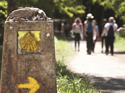 Yellow seashell signpost with small stones placed on top with people in distance walking along pathway for pilgrimage Way of St James, Camino de Santiago, Spain