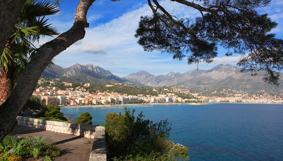 Distant view of French town Menton with tree overhanging at top of frame and sea between with mountainous landscape and blue sky with clouds in background, French Riviera, France