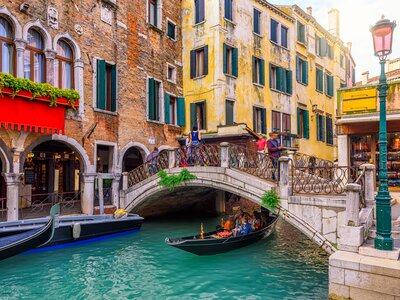 Narrow canal with gondola full of people passing beneath footbridge surrounded by colourful houses in Venice waterways, Italy