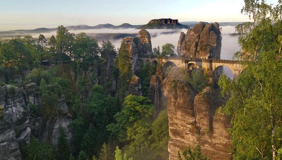 Bastei bridge with soft golden glow of sunlight casted over rocky peaks and trees with low clouds in distance, Saxon Switzerland National Park, Germany